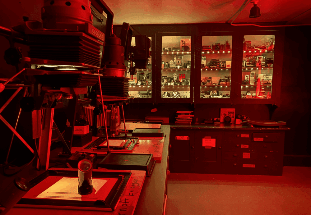 A dimly lit photography darkroom with red lighting, featuring multiple Beseler enlargers on a table. A cabinet in the background is filled with vintage cameras and photography equipment. The workspace is organised with various drawers and tools for developing photographs.
